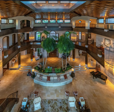 The inside of a mansion, with a centre room that is 3 stories in heigh, surrounded by landings for the other stories curving around outside of it. In the middle of the big room is 2 palm trees.