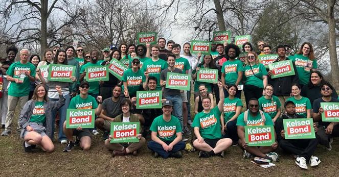 Dozens of people in green Kelsea Bond shirts gathered at a park holding signs, ready to go knock doors in the neighborhood and talk to people about voting.