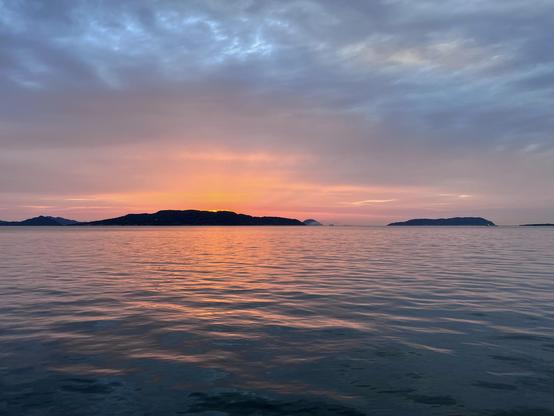 On the horizon line, a small island hides the setting sun while the sea reflects the warm pastel colors of the sky.