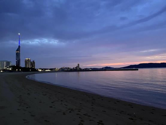The beach is getting darker but the buildings in the background are illuminated and the Fukuoka Tower spire is lit blue. Some pink lingers in the sky.