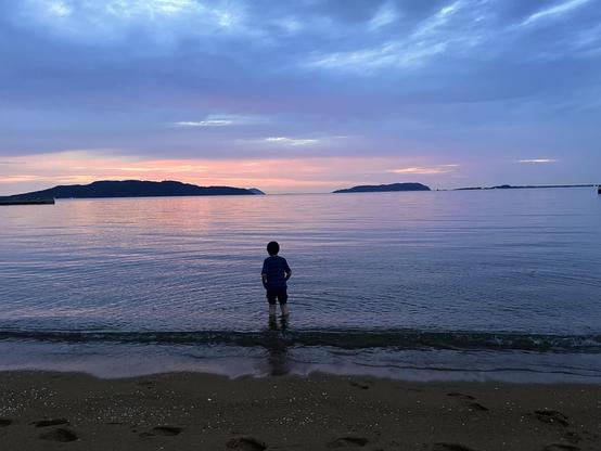A kid has walked into the sea up to his calves and is looking at the beautiful pastel colors of the sky.
