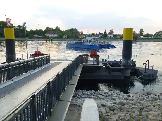 The image shows a waterway scene, likely near a dock or ferry terminal. There is a ramp leading down to the water, with yellow and black posts marking the area. In the background, a blue vessel is seen traveling on the water, and there are buildings and trees visible along the shoreline. The overall atmosphere appears calm, with the lighting suggesting either early morning or late afternoon.

Image Credits: Wikimedia / Muck / CC BY-SA 4.0