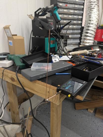 A photo of a J-shaped antenna made of copper wire. It is attached to a NanoVNA antenna testing gadget sitting on the edge of a bench. Lots of junk is visible in the background.