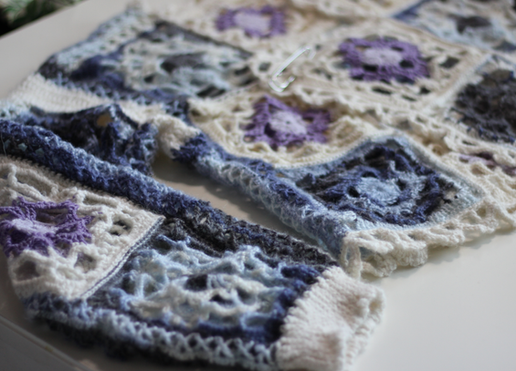 A crochet cardigan made in white, blue and purple yarns. It’s made up of grandma squares and edged with a little lace