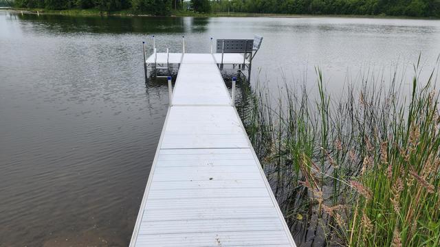 The image shows a modern, white aluminum dock extending into a calm body of water. The dock features a sturdy, metallic ladder with handrails on the left side, leading into the water. The surface of the dock is clean and smooth, with visible joints and supports. The water is still, reflecting the dock and the surrounding environment. The dock is supported by four vertical posts, each capped with a blue protective cover. The overall scene is serene, with no people or boats visible.