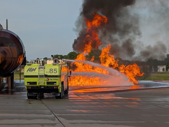 Aircraft simulator on fire for a simulated fuel spill with fire. The truck is on scene responding and managing the fire.