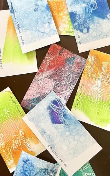 Business card-sized prints made using a gelli plate, acrylic paint and wood stamps. These are all different colours, some single colour, some many overlapping colours. Some motifs are visible; stamped, stylised leaves, flowers and a camel. The cards are scattered on a tabletop. These were pretty fun to make.