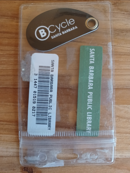 Photo  shows a Bcycle fob in a clear laminated badge holder, with Santa Barbara Public Library bar code and label.
The fob is curved with a flat bottom, brown background, white BCycle logo and "Santa Barbara" in white lettering.  There is a grommet at the right end, which would allow it to fit on a keyring.