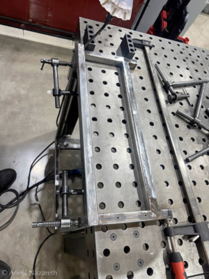 The top part of the frame is clamped to the table and across the frame, pulling two ends together. There is a metal bar spanning the frame