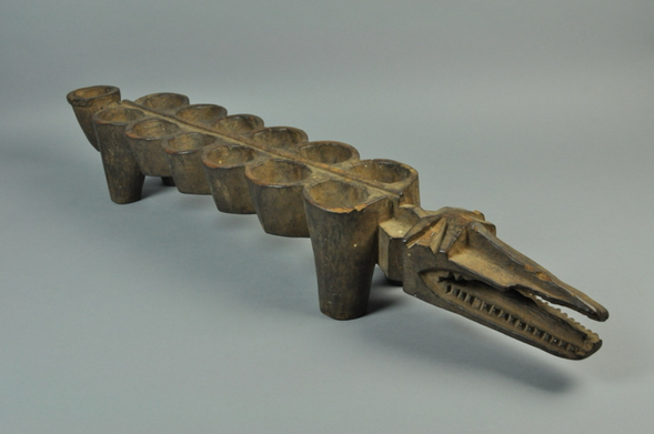 “Game board made of wood. Rectangular shaped scallop board supported by four legs. Projection in the shape of crocodile head at one end of board, opposite end has endcup projection. Twelve round playing pockets bisected by ridge.” Configuration: 2 x 6 Plus 1 Endcup (official museum photo, quarter turn side profile on light grey background)