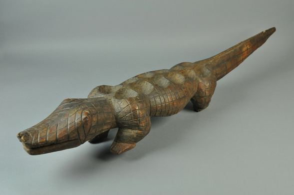 “Game board caved of wood in shape of a crocodile. Incised grid lines carved into entire figure to form the skin/scales of crocodile. Prominent snout, legs, and long tail. Twelve shallow playing cups carved into top/back of crocodile.” Configuration: 2 x 6 (official museum photo, quarter turn side profile on light grey background)