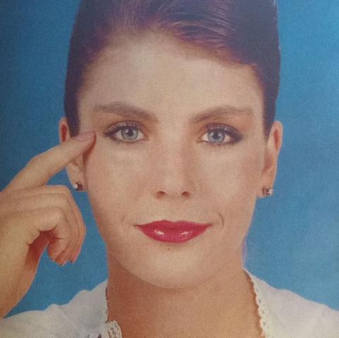 Retrato de Karen Sentíes  con cabello recogido, piel clara y labios pintados de rojo, señalando su ojo con un dedo mientras sonríe levemente. Fondo azul suave.