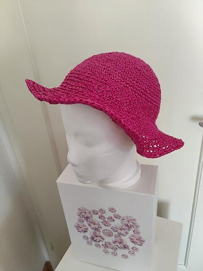 A pink crocheted sun hat made with paperyarn sits atop a white mannequin head. The mannequin is placed on a decorative white box featuring a floral design with pink flowers and a circular emblem. The background is a plain white wall.
