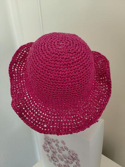 A bright pink crocheted sun hat with a wide brim is displayed on a decorative white box featuring a floral design. The textured, woven appearance of the hat is highlighted in this side-angle view. The background is a neutral, light-colored surface.