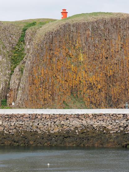 A bright orange lighthouse sits atop a grassy cliff, overlooking a rocky shoreline and calm water below. The cliff features textured, moss-covered rocks, and there are stones lining the water's edge.