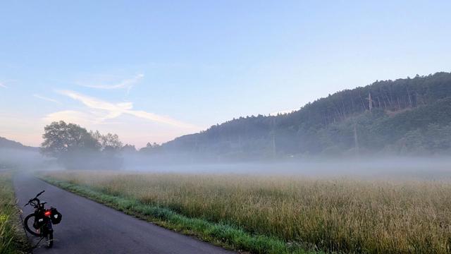 Ein ruhiges, weites Landschaftsbild im sanften Morgenlicht. Ein schmaler, asphaltierter Weg verläuft links durch das Bild. Ein Fahrrad mit Packtaschen steht am linken unteren Bildrand auf dem Weg, der von einem hohen Grasfeld gesäumt wird. Im Hintergrund liegt dichter Nebel oder Dunst, der sich über die Felder und am Fuße eines bewaldeten Hügels ausbreitet. Der Himmel ist hellblau mit einigen zarten Wolken.