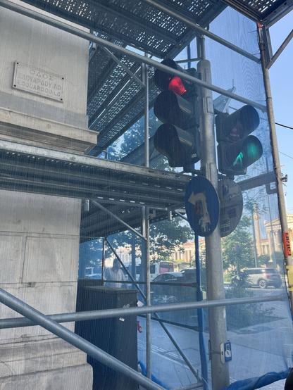 Looking past the marble corner of a building and through scaffolding and the taut blue material that covers it onto a busy avenue. Also behind the blue construction fabric, a traffic light shows red one way, green another. Across the avenue, the neoclassical Trilogy of University, Academy, and Library, are just in shot.