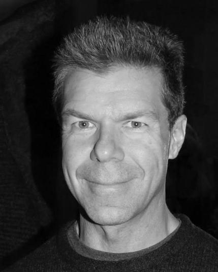 Black-and-white headshot of Gregory Norman Bossert, a white man with graying dark hair worn short and brushed straight up. He wears a crew-neck sweater over a lighter  tee. He looks straight into the camera and smiles a close-lipped smile that gives him laugh lines.