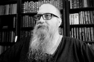 Head-and-shoulders black-and-white photo of writer David Anaxagoras, a fair-skinned man with a short hair and a long beard. He wears a dark woven shirt and black-framed glasses. He sits in front of a bookcase and looks into the distance with a serious expression.
