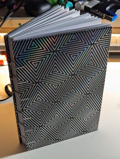 A coptic stitched notebook standing upright on a desk. The book is opened slightly, facing away from the camera to support standing upright. The cover is a holographic geometric pattern of triangle spirals on a black background. The book is bound with white thread that is visible on the front cover and across the spine.