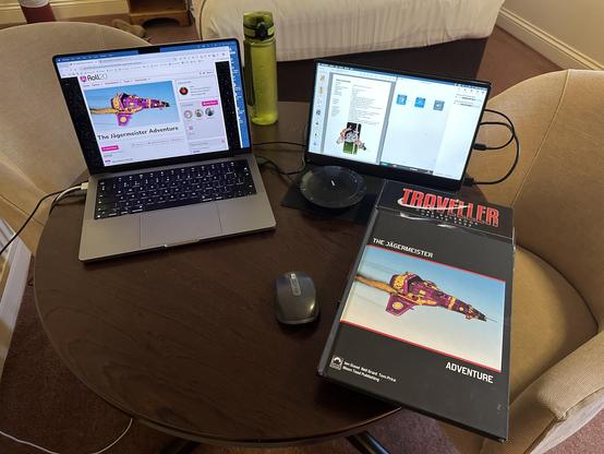 A hotel table with MacBook Pro and a portable second screen, mouse and conference speaker disk. Alongside is a copy of the Traveller core rules and the Jägermeister adventure. The computer screen has Roll20 open and the secondary screen has the scenario showing with AudioHijack ready to record the session.