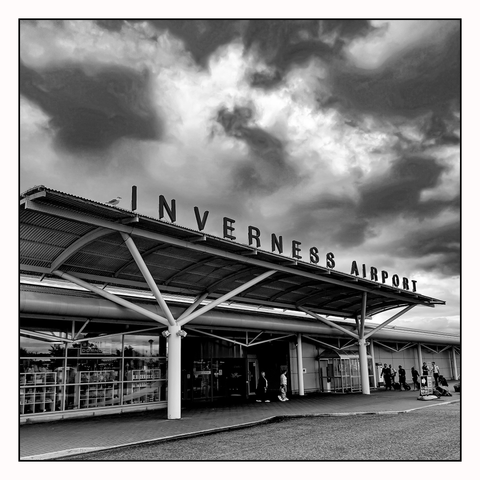 A modern looking building with tubular design, an overhanging section with large free standing letters spelling out ' INVERNESS AIRPORT' beneath a broiling moody sky.