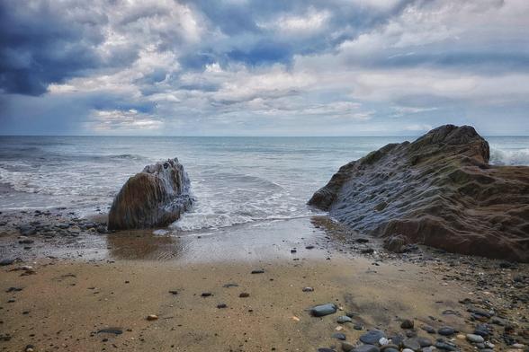 A dramatic coastal scene featuring two large, dark rocks on a pebble and sand beach. The ocean stretches out to the horizon under a heavily clouded sky. Waves gently lap against the shore and around the base of the rocks.
