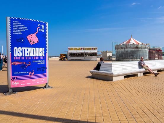 The image depicts a sunny day at a seaside promenade. In the foreground, a large blue and pink sign advertises the "Ostendaise North Sea Food Fest" scheduled for June 21-22, 2025, at Zeeheldenplein in Oostende.
In the background, a carousel with a red and white striped roof is visible, partially covered by a transparent fence. To the left, a food stall with a sign reading "Ice Cream Charros" is present. A couple of people are seated on white benches, enjoying the sunny weather. The sky is clear and blue, and the ground is paved with square tiles.
