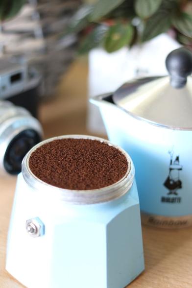 Bialetti moka pot with unscrewed top that is lying nearby. The filter of the bottom part is filled with coffee and in the background, there is a green plant