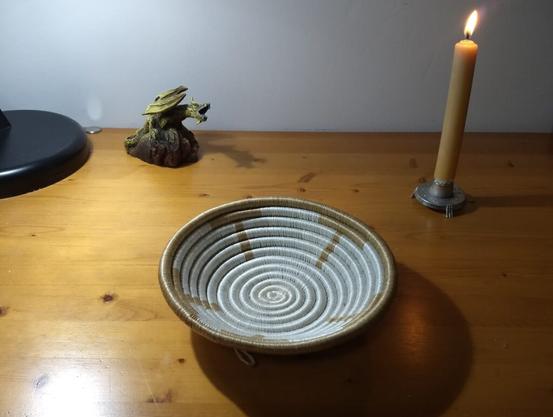 a coiled grass bowl wrapped with light brown and off-white fibres on a vanished wooden tabletop lit by a candle and an electric lamp