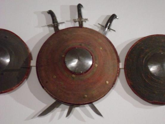 A round, domed wicker shield with a spiked steel boss and a cloth-bound rim. Three short scimilars hang behind it with their handles up and blades crossed at the middle.