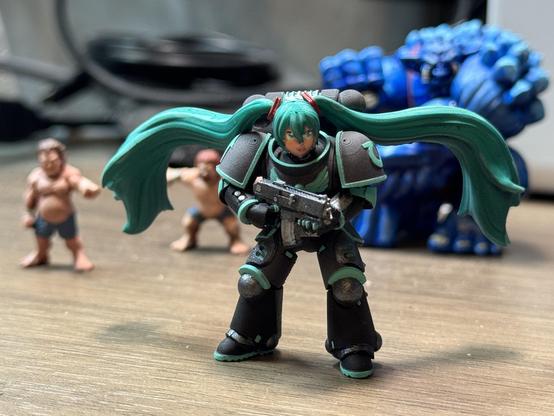 Space marine Miku from the front and in front of the out of focus Rumbleslam wrestlers in the background. One Halfling pointing a finger at her.