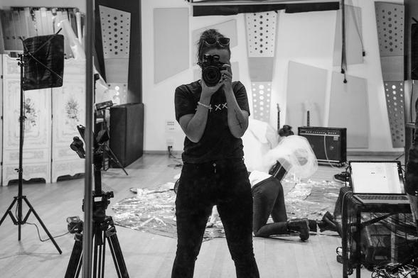 A black and white self portrait shot through a mirror in a rehearsal space. In the background a band can be seen setting up the room for a makeshift video shoot