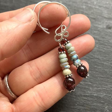 A photo of a pair of silver and stone bead dangle earrings held in the fingers of a hand. The earrings feature a tiny stack of stone beads (aquamarine & red wine garnet) with pewter accents hanging from hoop-shaped recycled sterling silver ear wires.