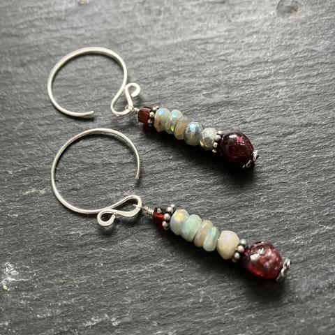 A photo of a pair of silver and stone bead dangle earrings on a dark grey slate background. The earrings feature a tiny stack of stone beads (aquamarine & red wine garnet) with pewter accents hanging from hoop-shaped recycled sterling silver ear wires. The angle of the shot highlights the way the stones glisten in the light.