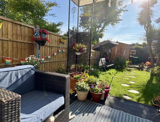View of my garden from a deck. We are under a black metal gazebo. There is an outdoor rug and a dark rattan chair with blue cushions. On the left is a fence adorned with baskets of flowers and rainbow bunting. The lawn to the right is green and the sky is blue. There are several pots of flowers on the deck. Two white cat legs peak out from behind the chair.