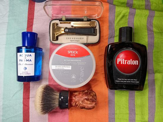 (Le Chat Mistral description)

The image displays a collection of grooming items arranged on a striped cloth. At the top left, there is a blue bottle of Acqua di Parma cologne. Next to it, an open clear case contains a gold-coloured Eversharp Hydro-Magic razor. Below the razor, there is a round tin of Speick Men shaving soap, which is vegan and dermatologically tested. In front of the shaving soap, there is a shaving brush with a wooden handle and dark bristles. On the right side of the image, there is a dark bottle of Pitralon aftershave. The items are neatly arranged, suggesting a well-organised grooming routine.