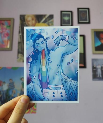 Hand holding up a print of my oc Isaiah flexing a bicep while holding a rainbow pride flag in his teeth, colorful confetti flying around.
