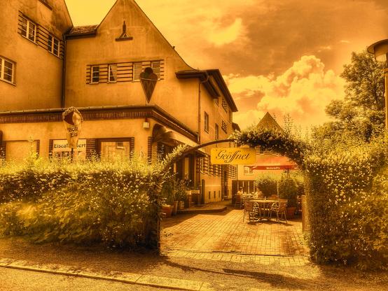 Bild in Amber. Eiscafé in Plauen. Links das Gebäude des Cafés, davor eine Hecke mit einem bogenförmigen Einlass, der den Blick auf die Terrasse freigibt. Der Himmel bewölkt.