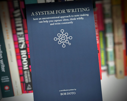 A book titled A System for Writing by Bob Doto is on display against a blurred background of other books.