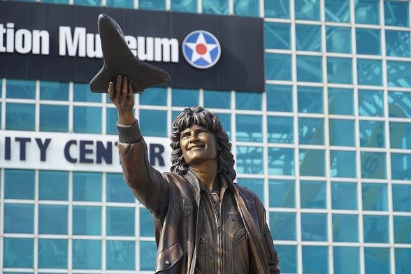 A bronze statue of a smiling woman in a flight jacket, holding a model of a space shuttle aloft. The background features a modern building with large glass windows and signs indicating an aviation museum and a community center.