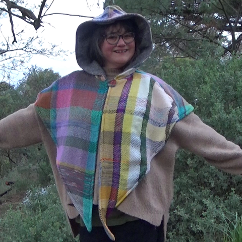 A white woman with dark hair and glasses, slightly chonky. It's me! I'm standing outside in front of some sketchy greenery. I'm wearing a capelet with a big hood and two long pointed panels at the front. The fabric is handwoven from scraps, so it is a mix of colours and stripe widths.