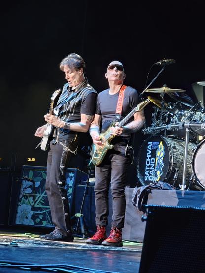 Steve Vai and Joe Satriani vibing, standing closely together playing guitar on stage.