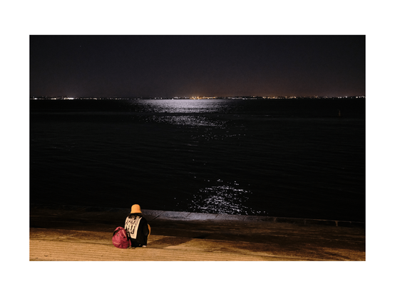 lonesome person from the back sitting at the riverside of tejo river in lisbon at night, staring over the water, moonlight reflexions in black water