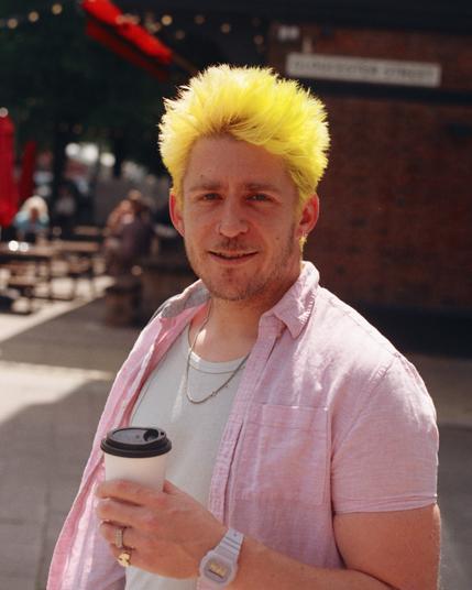 A masc person with bright green/yellow hair, pink over shirt, white undershirt, holding a coffee, looking at the camera