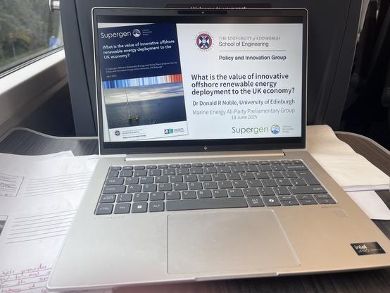 A laptop on a train table, with the opening slide of a presentation. There is a picture of a report and the title:

What is the value of innovative offshore
renewable energy deployment to the
UK economy?

Dr Donald R Noble, University of Edinburgh
Marine Energy All-Party Parliamentary Group
18 June 2025

At the top is the logo for The University of Edinburgh, School of Engineering, 
Policy and Innovation Group and at the bottom a logo for the Supergen Offshore Renewable Energy hub