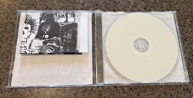 The image shows a partially open CD case featuring a blank CD on the right side and a small booklet with black and white artwork on the left side. The case is placed on a textured surface.