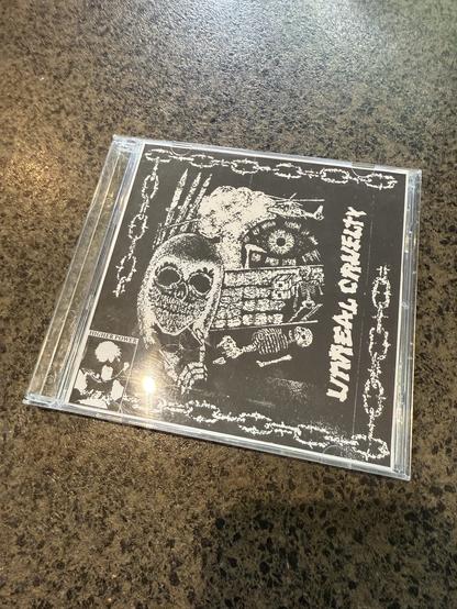 An album cover featuring a black and white design with a skull, skeleton imagery, and graphic elements. The title "Unreal CRUELTY" is displayed prominently. The cover is placed on a textured surface.