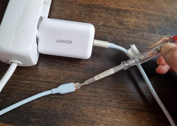 A photo of a white Anker USB-C charger plugged into a white, 3-slot power strip lying on top of a dark wooden table. Plugged into the charger is a light blue cable which after going out-of-picture returns into view with the USB-C plug being in the centre of the image. Held up to the plug is a dimly glowing phase tester screwdriver.