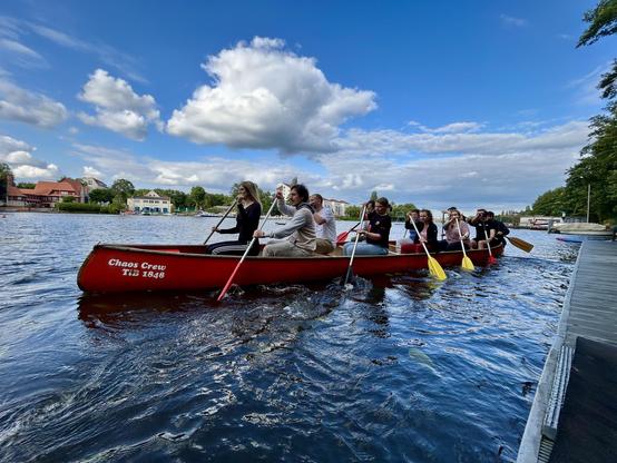 Ein Team paddelt gemeinsam in einem roten Drachenboot mit dem Schriftzug „Chaos Crew“ auf einem Fluss, bei sonnigem Wetter und unter blauem Himmel.